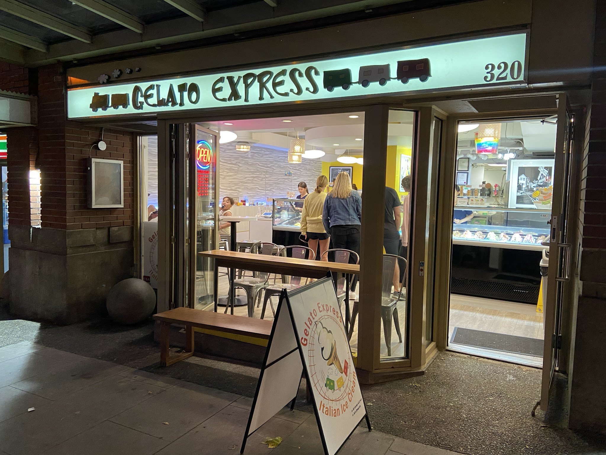 Gelato Express storefront at night showing illuminated sign, outdoor bench seating, and customers visible through the open door